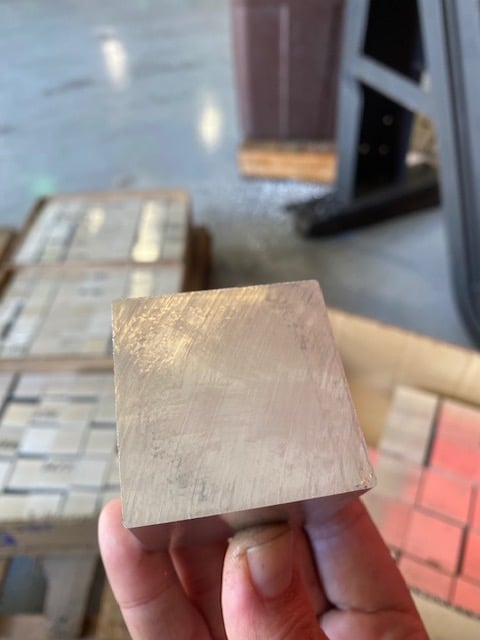 Hand holding a light-colored wooden block or sample with construction materials and flooring visible in the background
